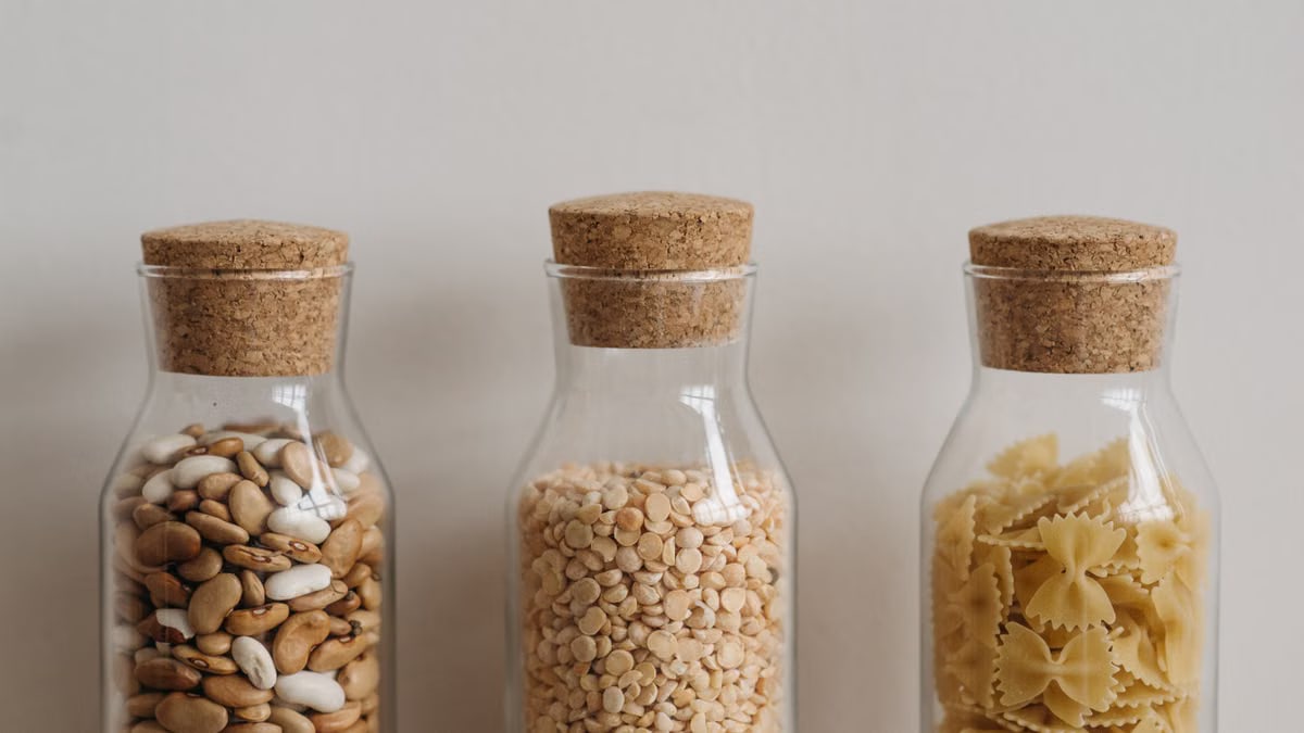 Three clear glass jars with cork lids hold beans, lentils, and pasta showcasing pantry organization.
